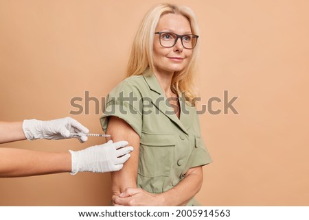 An adult caucasian beautiful blond woman in glasses comes to have an injection, squeezes her elbow nervously, tries to stay positive. Nurses' hands in white medical gloves hold the injection site. 