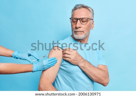 Protect yourself from coronavirus with vaccination. Serious bearded grey haired man gets injection in arm for staying safe wears spectacles t shirt isolated on blue background. Treatment for covid 19