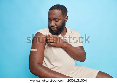 Serious dark skinned man looks at arm with plaster got vaccinated during coronavirus panemic sits indoor against bluebackground after vaccine injection shows shoukder with bandage. Immunization