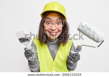 Positive glad Asian construction worker dressed in uniform holds painting brush and roller wears protective transparent glasses uniform isolated over white background. Refurbishment home improvement