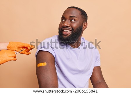Health care global vaccination of population. Handsome cheerful dark skinned bearded man looks away hears consultation from doctor gets vaccine against coronavirus poses against brown background