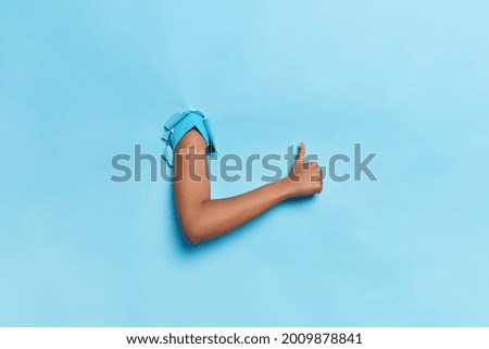 Unknown person breaks arm through paper background gestures against blue wall keeps thumb up makes like gesture recommends to do vaccination wears adhesive plaster. Population immunization concept