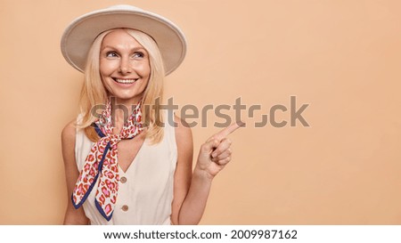 Glad forty years old woman in stylish hat dress and kerchief on neck indicates away on copy space advertises something shows way to shopping mall isolated over brown background. Place your promo here