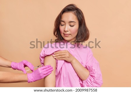 Health care and inoculation concept. Brunette Asian woman looks at process of vaccination gets antiviral vaccine to reduce covid 19 spread wears in stylish clothes. Unknown nurse makes injection