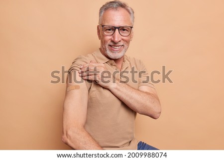 Senior man in spectacles shows plastered arm after getting coronavirus vaccine happy to feel safe and protected isolated over brown background. Health care during pandemic. Old people vaccination
