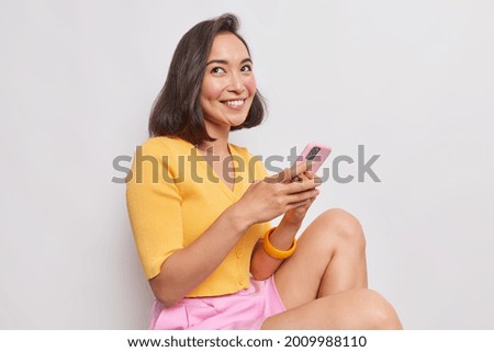 Pretty young Asian woman with bob hairstyle smiles gently has dreamy expression sits against white background holds mobile phone sends text messages wears yelow jumper pink shorts feels happy
