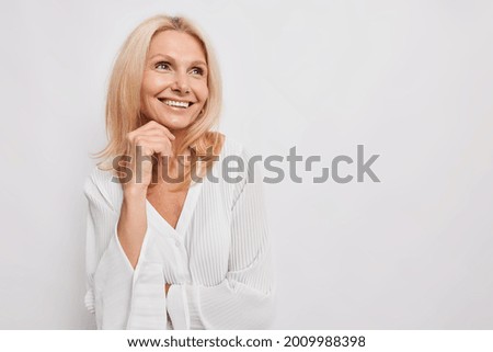 Beautiful young European woman of middle age smiles gently keeps hand under chin looks away with dreamy expression wears silk blouse isolated over white background copy space for advertisement