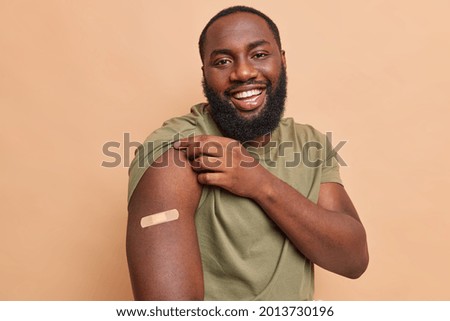 Cheerful dark skinned man shows adhesive plaster on shoulder after getting coronavirus vaccine feels safe gets injection in arm cares about health during pandemic isolated over beige background