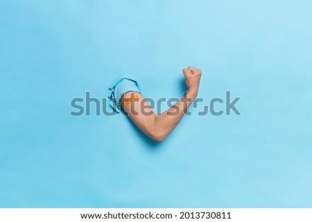 Horizontal shot of unrecognizable human keeps arms in paper hole clenches fist demonstrates power and strength after vaccination feels protected isolated over blue background shows bandage after shot