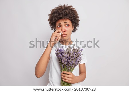 Unhappy ethnic woman with curly hair has inflammation of eyes runny nose sprays nasal aerosol being allergic to lavender looks sadly somewhere poses against white background. Allergy symptoms