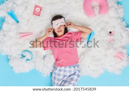 Pleased brunette Asian woman awakes after healthy sleep wears sleepmask on eyes dressed in pajama poses on white fluffy cloud surrounded by different items. Good morning and awakening concept