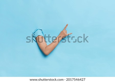 Faceless person shows arm with adhesive plaster makes rock n roll gesture get vaccine shot in shoulder isolated over blue background. Antiviral program for population against coronavirus pandemic
