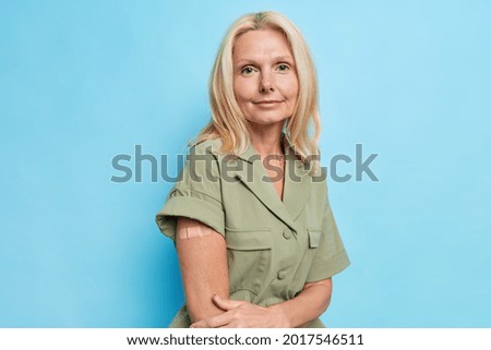 Serious blonde European woman shows vaccinated arm after vaccine injection wears dess poses against blue background looks directly at camera. Health protection immunization and safety concept