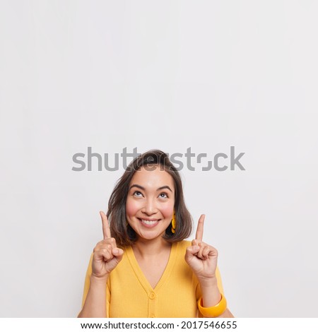 Positive brunette Asian woman points above shows place for your advertising text smiles pleasantly wears yellow jumper earrings and bracelet isolated over white background. Great idea cool offer