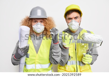 Indoor shot of professional maintenance workers wear protective helmets safety respirators uniform hold blueprint and painting roller inspect construction site work on house decoration and improvement