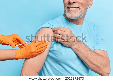 Health care concept. Unknown elderly man gets coronavirus vaccine shot in shoulder wears casual t shirt smiles toothily isolated over blue background. Medical doctor or nurse makes injection