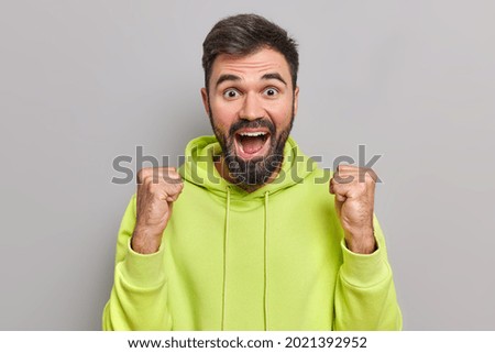 Cheerful bearded handsome man raises fists in victory gesture celebrates success or triumph exclaims from big joy wears casual green sweatshirt isolated over grey background. Yes I did it finally
