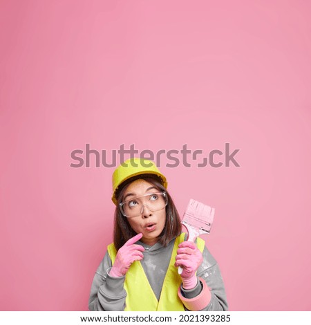 Interior color trends. Surprised Asian woman focused away thinks about apartment refurbishment holds painting brush wears uniform protective helmet isolated over pink background blank space.