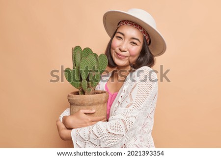 Portrait of young Asian woman embraces potted cactus wears fedora white knitted jumper has rouge cheeks natural beauty isolated over beige background. Houseplants concept. Botany and people.