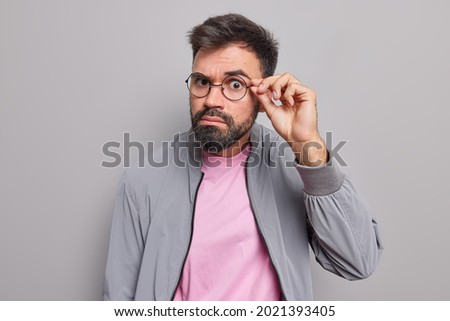 Serious bearded man keeps hand on rim of spectacles has attentive gaze directly at camera has surprised expression wears jacket isolated over grey background listens something. Monochrome shot