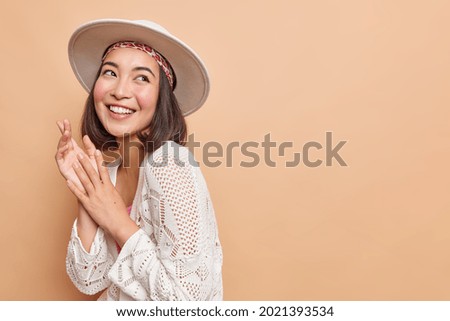 Prettty smiling Asian woman with dark hair expresses positive authentic emotions looks gladfully aside dresed in fashionable clothes isolated over beige background copy space for your information