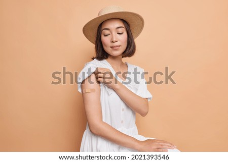 I got my coronavirus vaccine. Serious Asian lady looks attentively on place of inoculation wears aid band got antiviral injection for coronavirus protection wears white dress and fedora poses indoor
