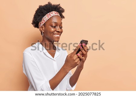 Sideways shot of dark skinned woman holds modern mobile phone surfs net checks newsfeed in social networks wears headband white shirt isolated over beige background copy space for promotion. 商業照片 © 