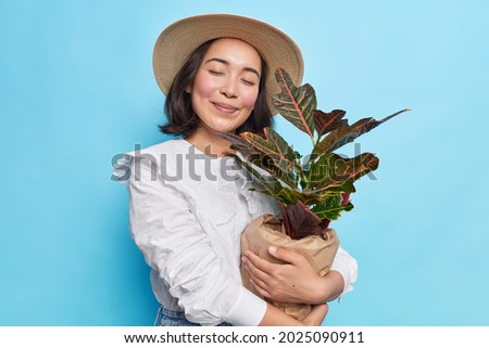 Pleased young Asian woman closes eyes embraces pot of houseplant happy to buy new flower for her home garden wears hat white blouse isolated over blue background. People and lifestyle concept