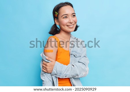 Pleased teenage girl with eastern appearance has two pony tails smiles gently happy to get vaccine against coronavirus dressed in orange dress and denim jacket isolated over blue background.