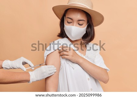 Horizontal shot of Asian woman wears protective sterile mask to prevent coronavirus spread gets vaccine injection in shoulder poses against beige background. Health care immunization and vaccination