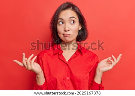 Clueless hesitant beautiful Asian woman with short dark hair spreads palms sideways shrugs shoulders has indecisive expression dressed in shirt stands against vivid red background. Who knows. Foto stock © 
