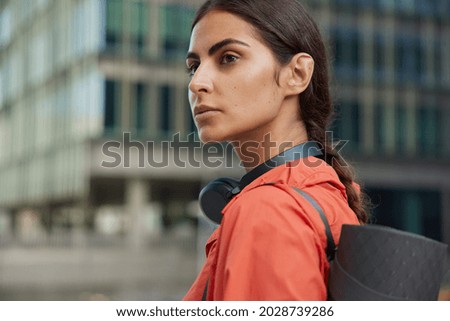 Sideways shot of pensive brunette young woman looks away into distance wears headphones around neck carries karemat strolls in city outdoors returns home from workout leads active lifestyle.
