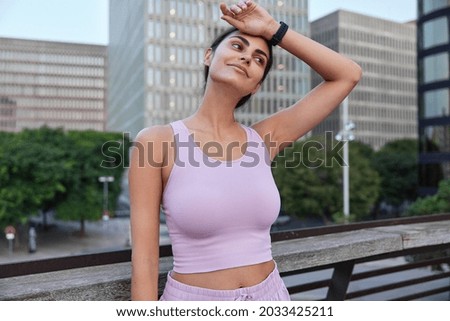 Tired sportswoman wipes forehead after hard fitness training wears cropped top smartwatch looks thoughtfully forward poses at bridge against tall cityscrapers view being in good physical shape