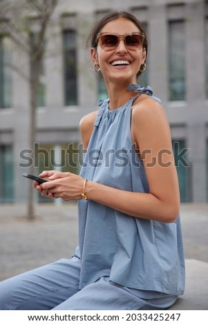 Optimistic glad woman with dark hair white perfect teeth uses mobile phone surfs social networks wears sunglasses and blue summer costume sits outdoor against urban background chats with friend