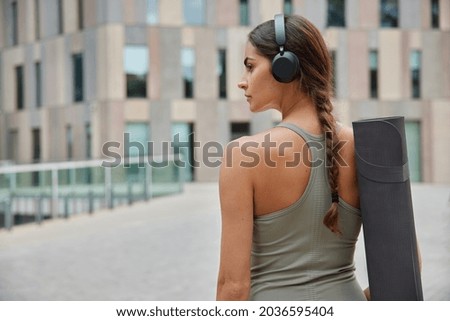 Healthy active lifestyle. Back view of sportive woman works out outdoors ready practice yoga does sports training or stretching walks with karemat listens popular music playlist goes to sport center