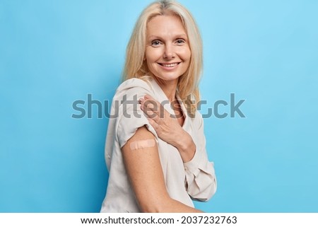 Sideways shot of blonde forty years old woman approves immunization campaign gets inoculation from covid 19 virus feels protected isolated over blue background. Vaccinated middle aged European lady