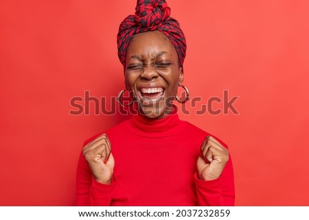 Excited beautiful dark skinned woman clenches fists as sign of victory celebrates success wins lottery keeps eyes closed from satisfaction wears turtleneck and silk scarf on head isolated on red wall