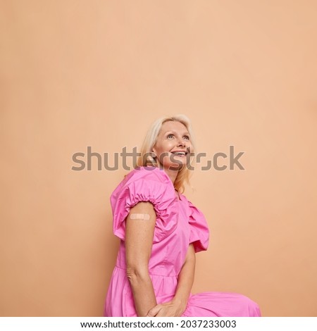 Health protection vaccination of population. Glad middle aged blonde woman shows shoulder with band aid after getting vaccine involved in immunization against coronavirus beige studio background.