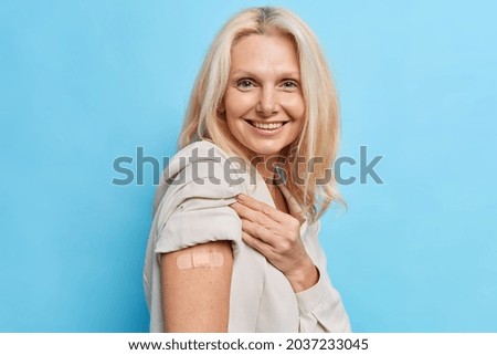 Glad woman shows shoulder with band aid after injection cares about health happy after getting vaccine shot poses against blue studio wall. Disease preventionn coronavirus immunization concept