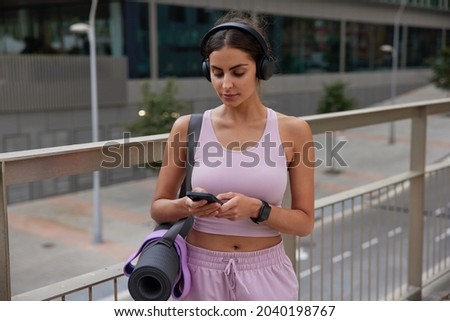 Woman does sport activities programes fit watch synchronizing data with mobile phone wirelessly checks health application enjoys audio song in headphones exercises outdoors to boost metabolism rate