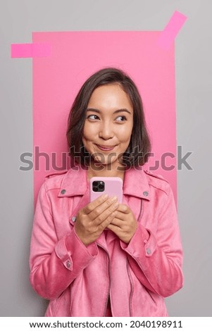 Lovely dreamy young Asian woman holds mobile phone chats with friends uses cool gadget and application dressed in jacket poses against grey background with plastered pink paper for your info