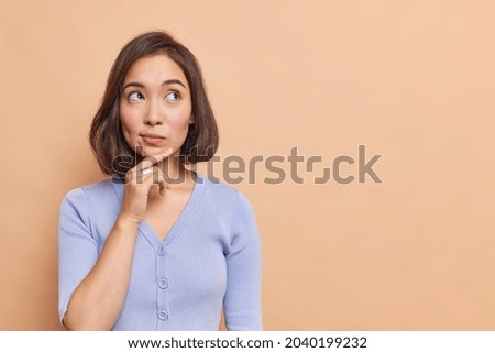 Photo of Thoughtful Asian woman keeps hand on chin looks pensively above dressed in casual blue jumper poses against brown background blank copy space for your advertising content thinks about future