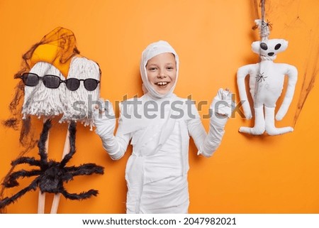Autumn traditional holiday of all saints. Positive small female child pretends being mummy or zombie prepares for halloween celebration isolated over orange background. Magic and miracles concept