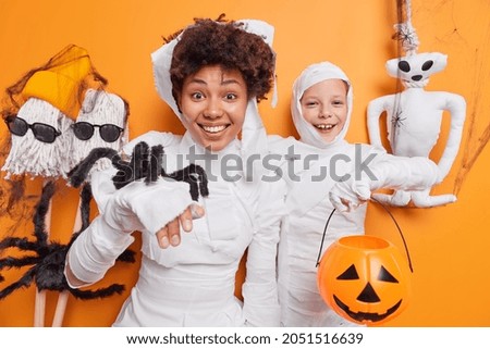 African American woman and small child wear halloween costumes hold spider and carved pumpkin jack o lantern pose against orange background with spooky handmade toys around. Happy celebration