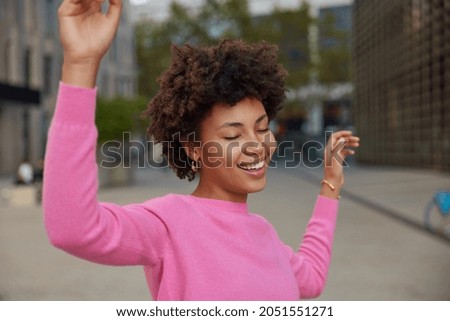 Glad relaxed young African American woman with curly hair dances carefree keeps arms raised dressed in casual pink poloneck closes eyes from satisfaction poses against blurred city background