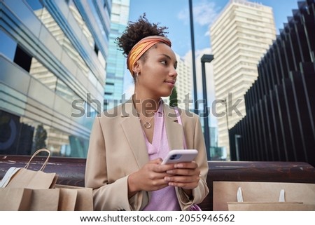 Young fashionable Afro American woman shopper satisfied after making purchases sits on wooden bench outdoors in center of city surrounded by paper shopping bags uses cellphone for networking