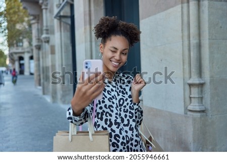 Happy female shopper with two combed buns poses with paper shopping bags enjoys fashion sale carries shop purchases makes selfie on modern smartphone winks eye at camera of cellular. Shopaholic