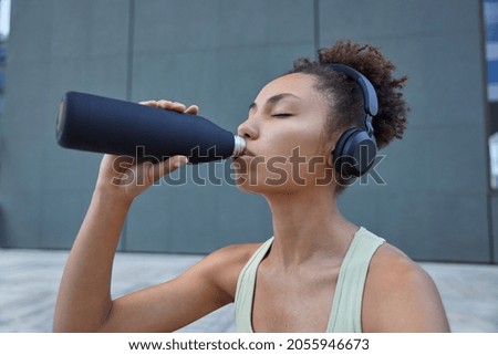Sweaty tired young sportswoman with curly combed hair drinks refreshing water from bottle feels thirsty listens music in headphones poses outside against dark background rests after fitness exercises