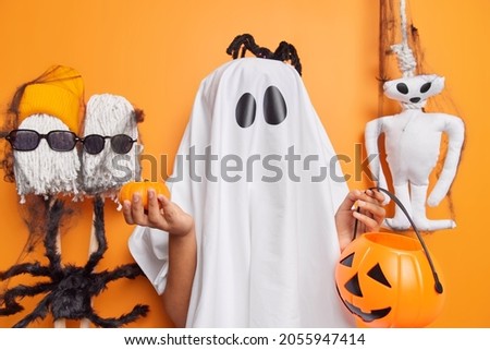 Horizontal shot of scary creature dresses up in halloween costume covered with white sheet holds two pumpkins has black spider on head poses against orange background. People mystery spooky event.