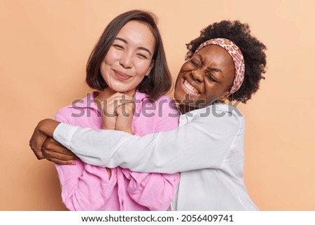 Friendly women embrace each other have good relationships dressed in casual clothes smile gently isolated over beige studio wall. American woman cuddles her best friend missed her